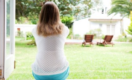 A woman sits at an open doorway with her back to the camera. She is facing a garden with a green lawn, trees and wooden chairs.